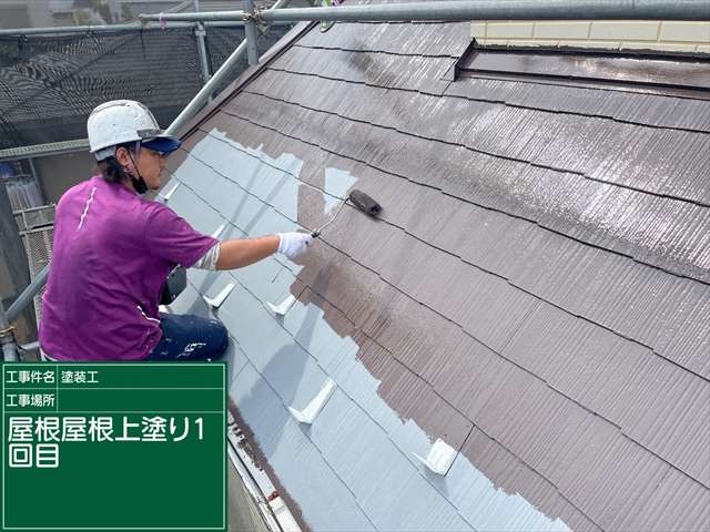 下塗りで白くなった屋根が紫の服を着て白いヘルメットを被った職人の手によって茶色く塗られていく様子。